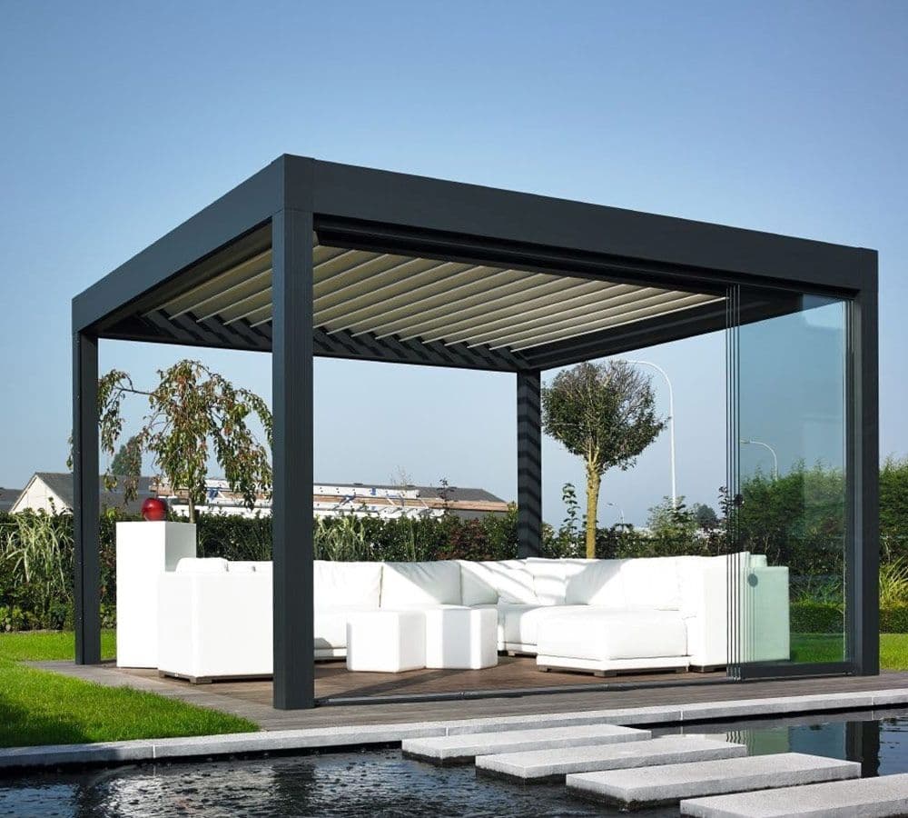Antrasit renkli biyoklimatik pergola; yan tarafı sürme cam ile kapatılmış, altında beyaz oturma grubu.