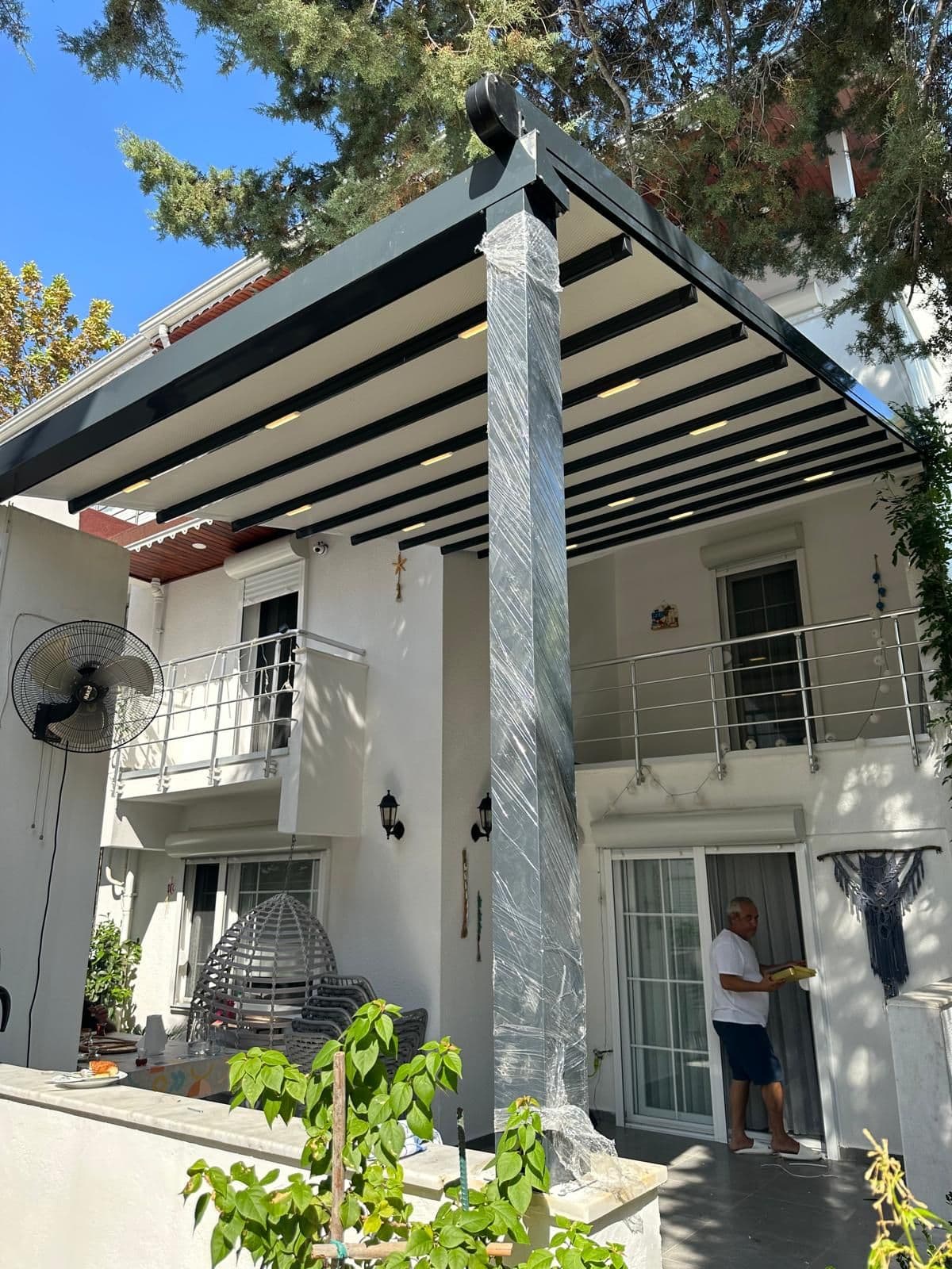 Villa cephesine monte edilmiş antrasit renkli motorlu pergola tente; çatı örtüsü açık konumda ve profillerde gömme LED aydınlatma.