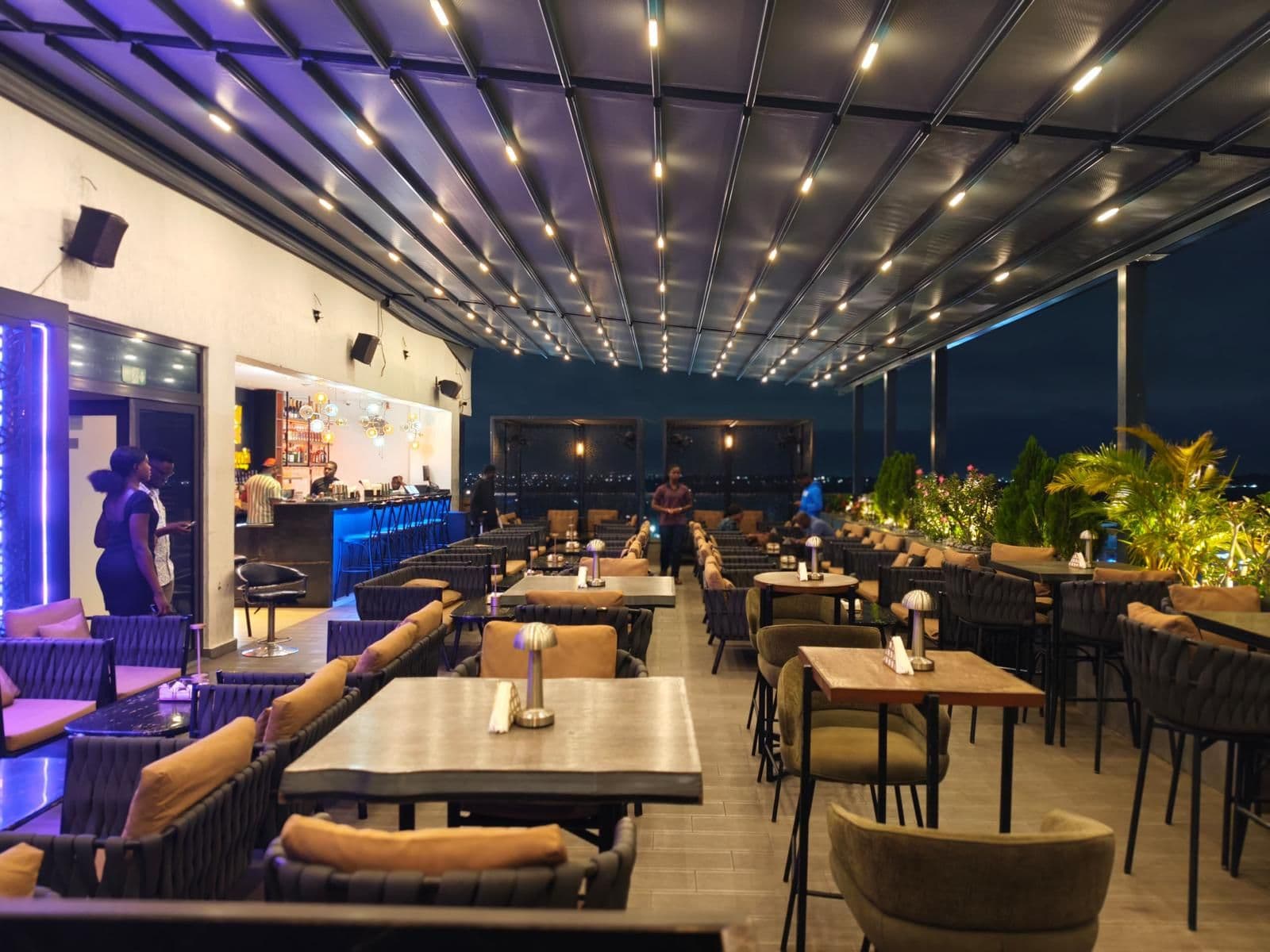 Gece açık olan bir restoran terası; üstte LED aydınlatmalı motorlu tente sistemi, altında dolu oturma alanı.